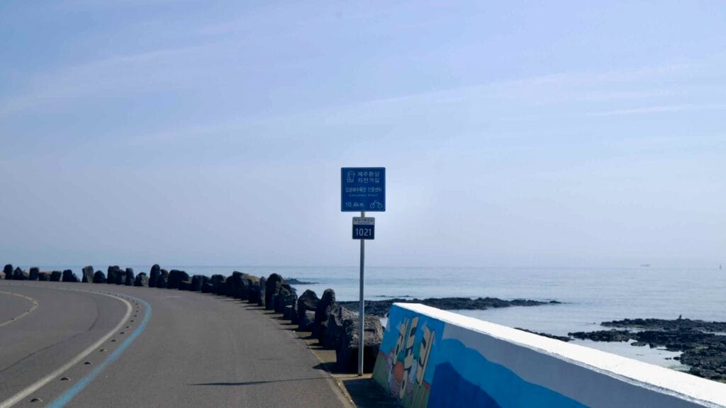 A Jeju Fantasy Bike Path sign marks the coastal route near Handong with a seawall mural and calm sea.