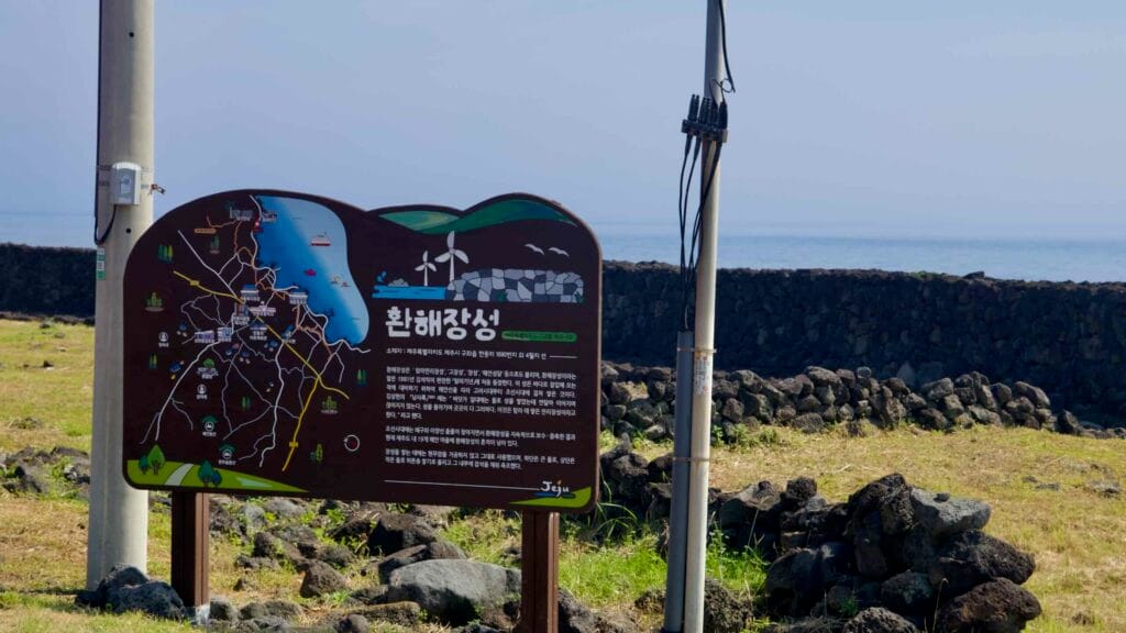 A Hwanhaejangseong information sign stands before long basalt field walls near Handong.
