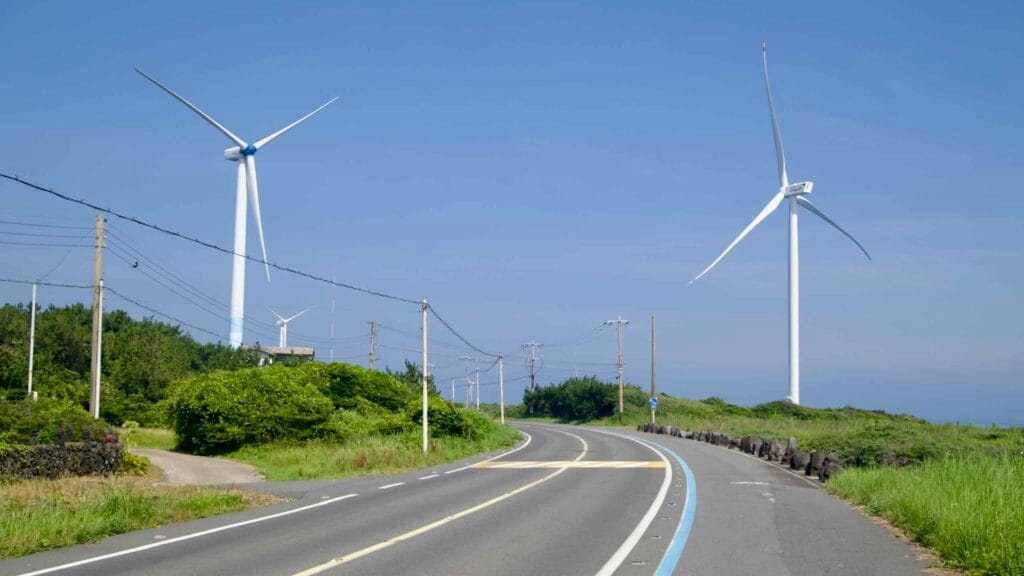 A coastal road with a bike lane curves through Haengwon-ri beneath tall wind turbines.