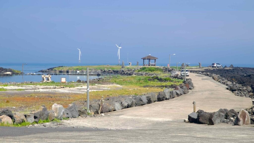 A concrete causeway reaches a pavilion at Haengwon Port with offshore wind turbines beyond.