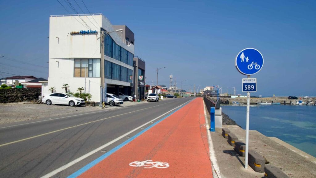 An orange coastal bike lane crosses Haengwon Port’s bridge past shops and the harbor.