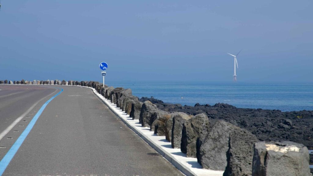 Basalt bollards line a coastal curve at Haengwon with an offshore wind turbine on the horizon.