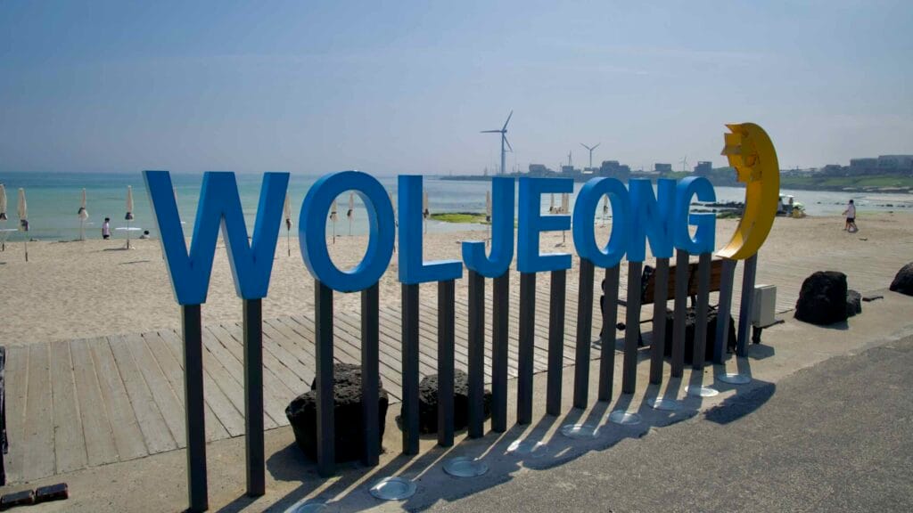 Blue “WOLJEONG” letters stand on the sand with turbines beyond.