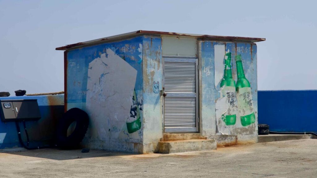 A weathered port shed shows peeling soju murals on the breakwater.