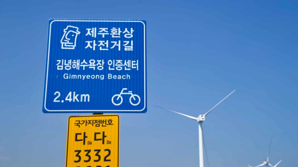 A Gimnyeong cycling sign stands with turbines beyond a clear sky.