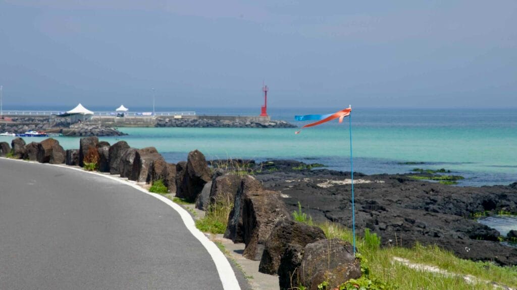 Gimnyeong’s coast curves past lava-stone guards toward Segial’s red lighthouse.
