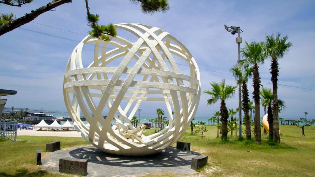 A white interlaced sphere sculpture stands on a park terrace above Hamdeok Seoubong Beach.