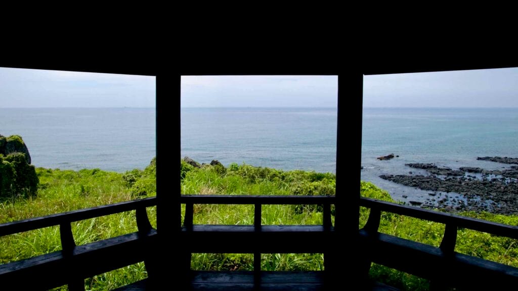 Dark gazebo on a hilltop overlooks the East Sea, with green slopes and a lava reef below.
