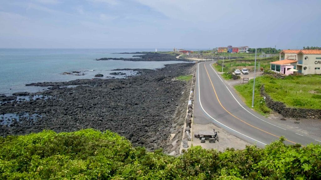From Shinheung hillside, the coast road winds past black lava reefs to a white lighthouse.