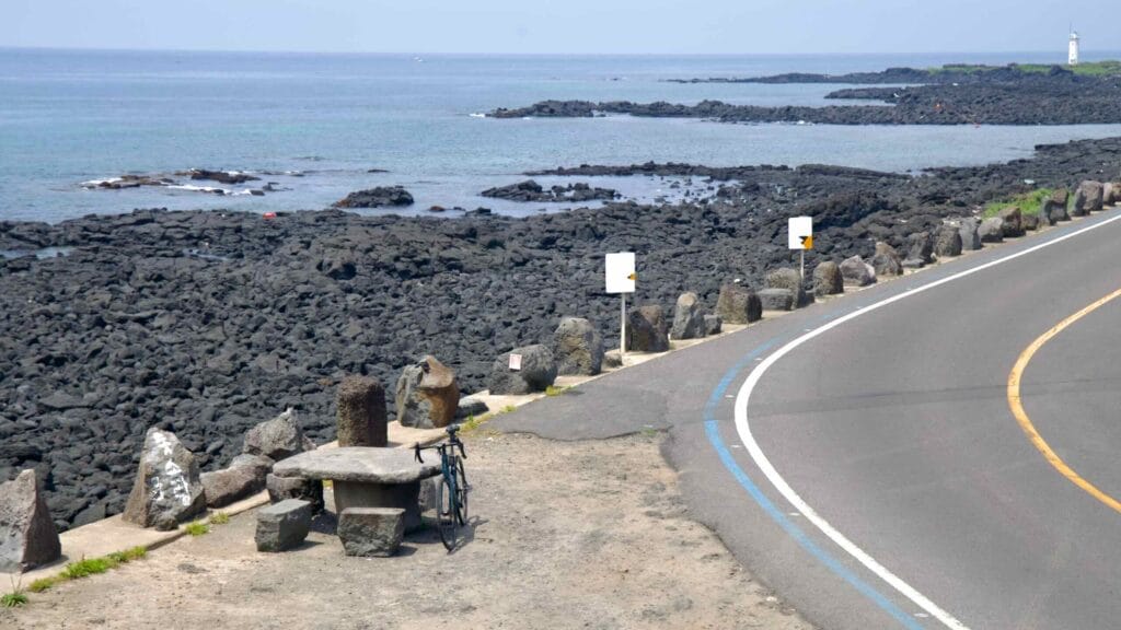 Bike by stone table near Jocheon’s rocky shore, blue bike lane, and distant white lighthouse.