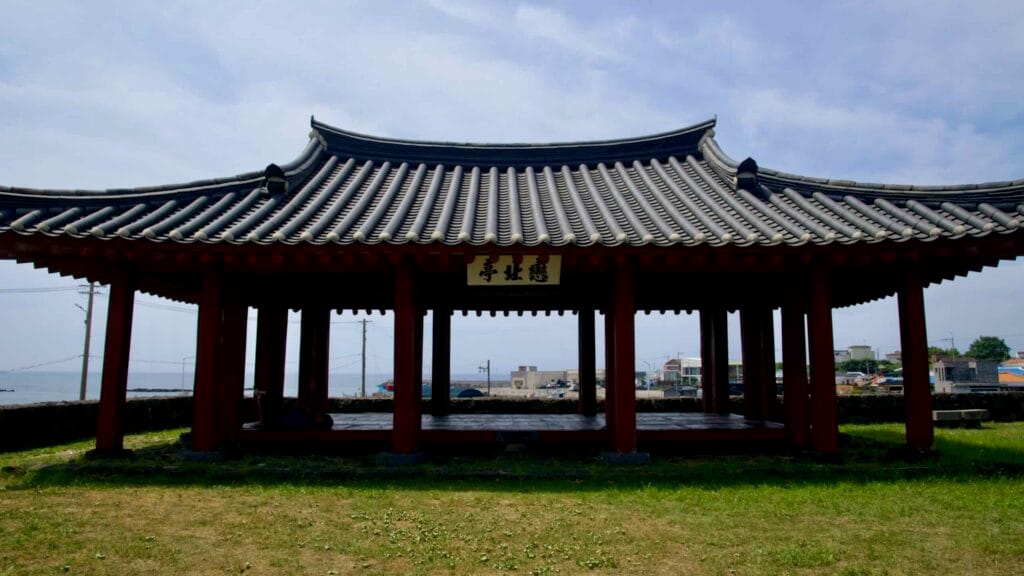 Under a tiled roof, Yeonbukjeong’s open hall overlooks Jocheon Port, framed by red posts.