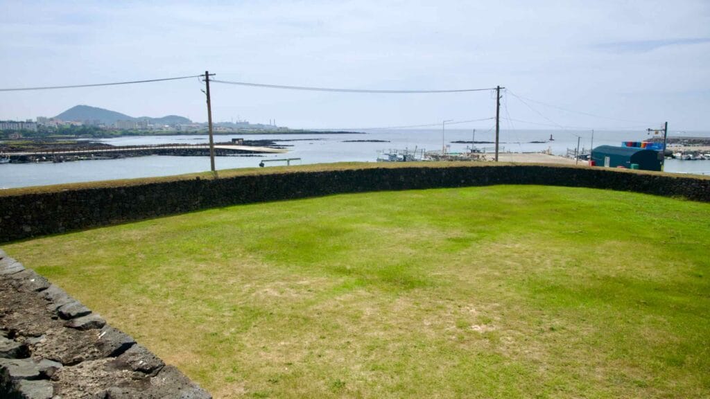 A trimmed grass yard below Yeonbukjeong overlooks Jocheon Port with boardwalks and fish traps.