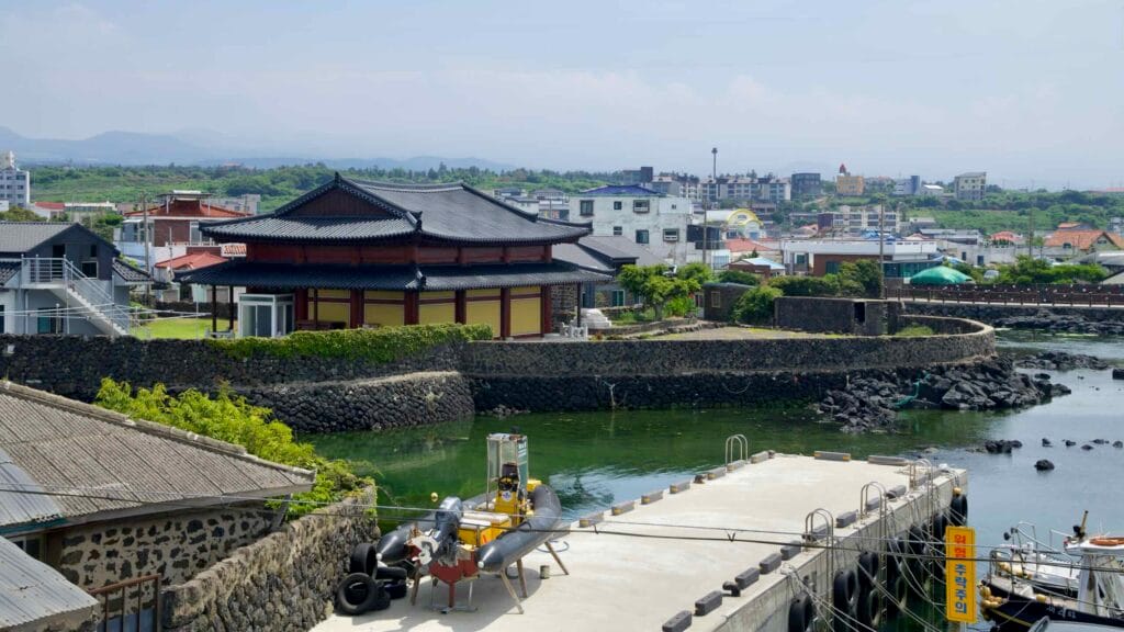 Traditional hall rises behind ivy-clad stone banks along green tidal water at Jocheon Port.