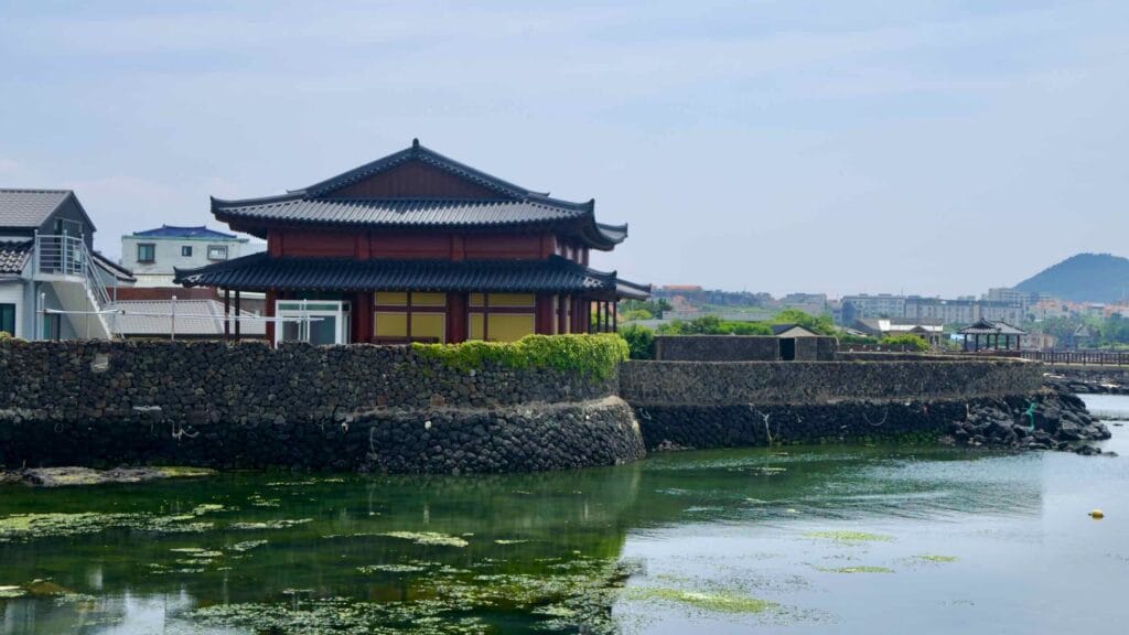 The waterside hall’s tiled roof reflects on the calm inlet at Jocheon beneath low neighborhoods.