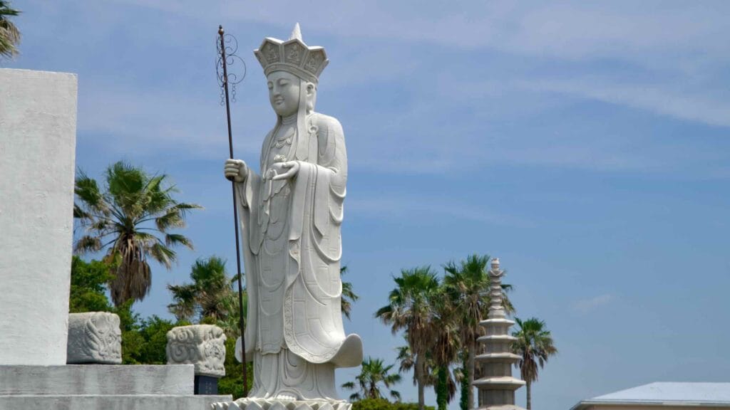 A standing Bodhisattva holds a staff beside a stone pagoda and palms.