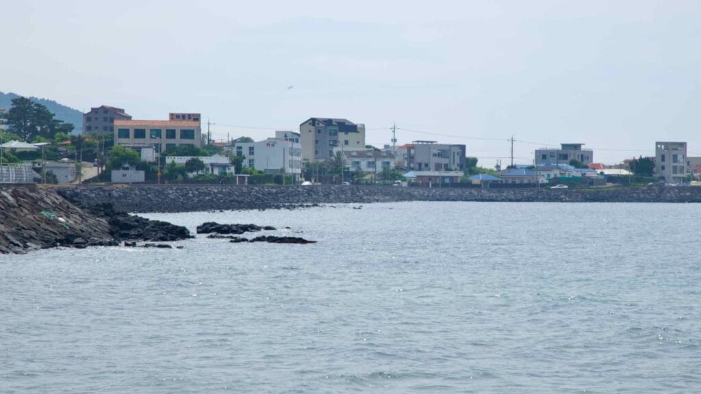 Low houses line Samyang-dong shore beyond a rock seawall as the East Sea laps basalt outcrops.
