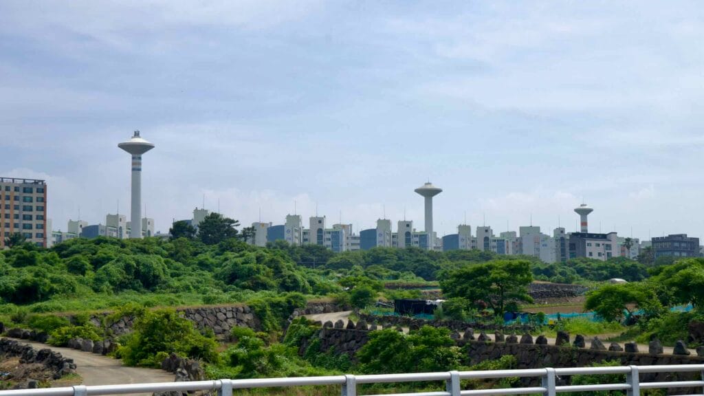 Water towers and apartments rise behind green belts and stone walls east of Jeju Harbor.