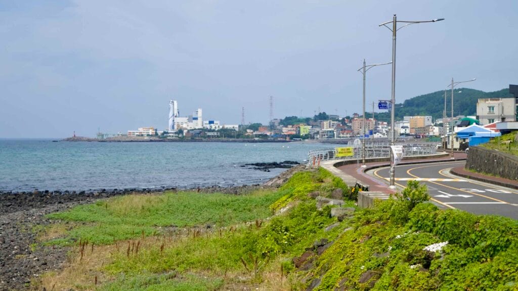 Samyang–Hwabuk coast road curves to an industrial shore with red lighthouse and rocky tidal flats.