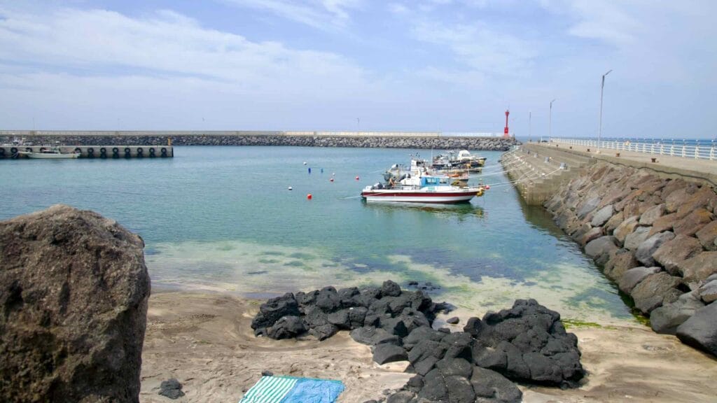 Small fishing boats float in the sheltered basin of Beollang Port behind high breakwaters and a red lighthouse.