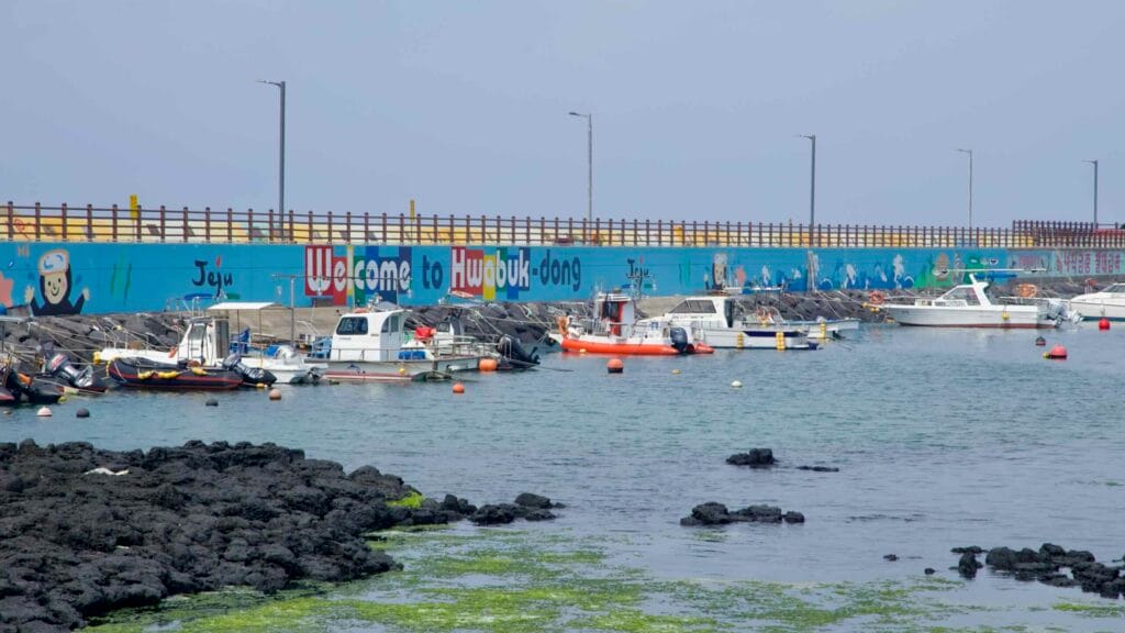 A close view shows the colorful "Welcome to Hwabuk-dong" seawall with moored boats.