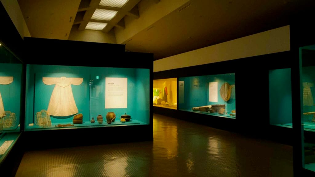 Glass cases display garments, pottery, and tools, with lit panels and a corridor of exhibits in the gallery.