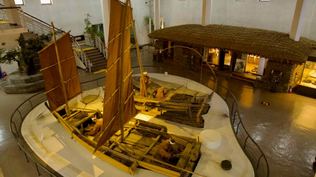 An overhead view shows wooden sailing craft and a thatched farmhouse installation in the museum.