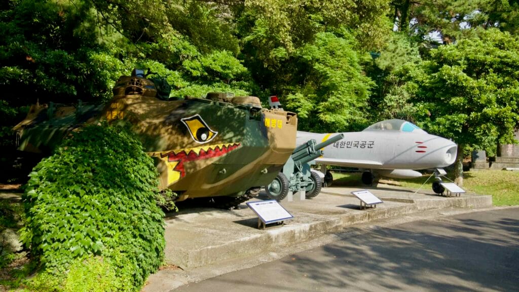 Armored vehicle and a small jet fighter stand on outdoor plinths at Jeju Unification Hall.