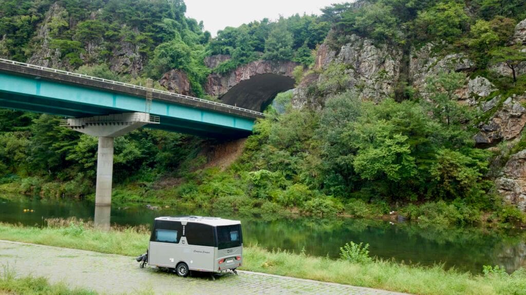 A bridge crosses a calm river beside a rock-cut tunnel and a parked camper.