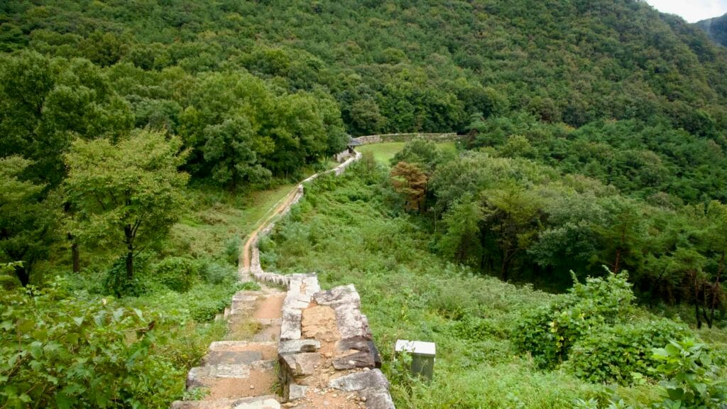 A rebuilt wall-walk and steep steps drop toward a small gate pavilion in the forest.