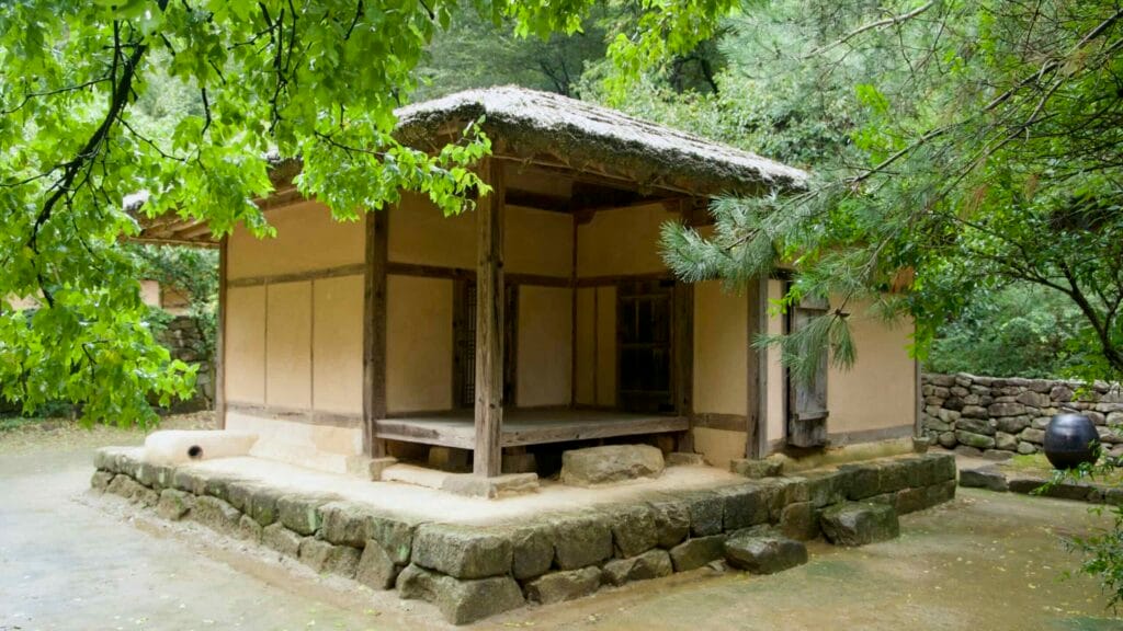 A small thatched house stands on a stone base in the forest near the old road.