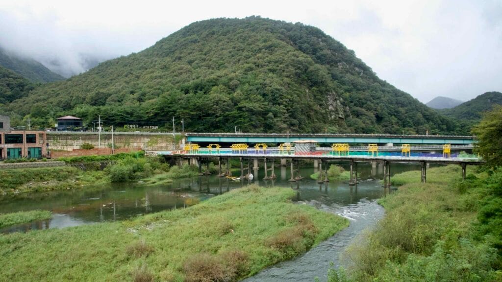 Colorful rail bikes and service bridges cross the Namhangang below wooded hills, with nearby buildings and roads along the riverbank.