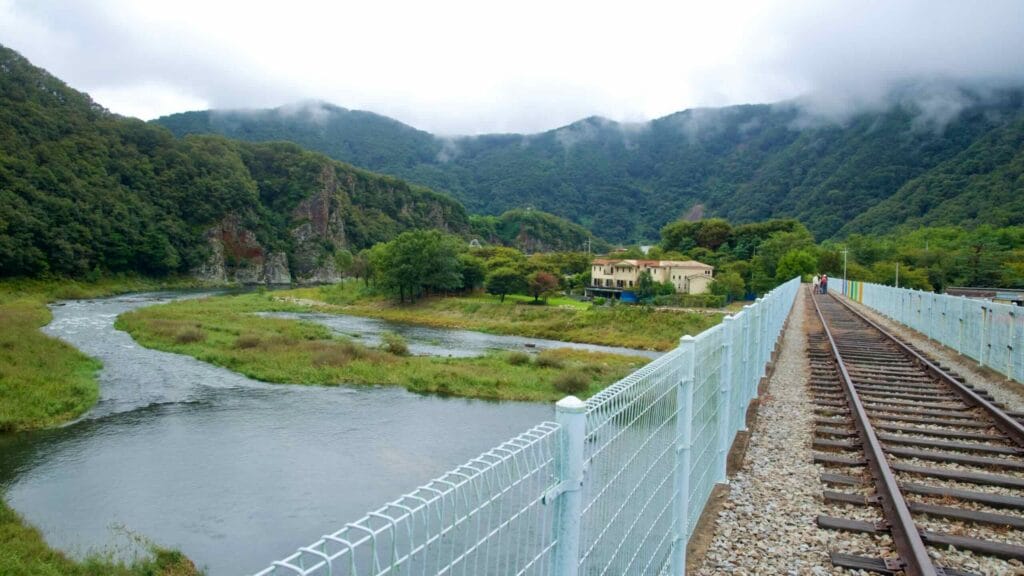 From the rail bridge, the Namhangang curves past villas and trees toward sheer rock cliffs at Jinnamgyoban, while fog drapes the ridges.
