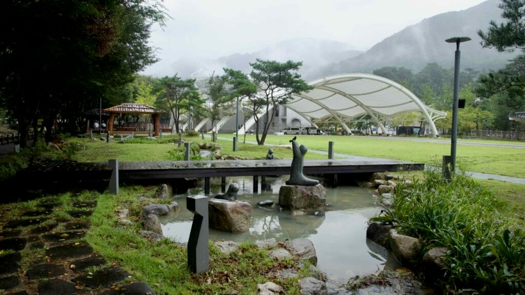 A narrow stream and wooden footbridge lead to lawn sculptures and a white tensile pavilion at Mungyeong Saejae with gazebos nearby.