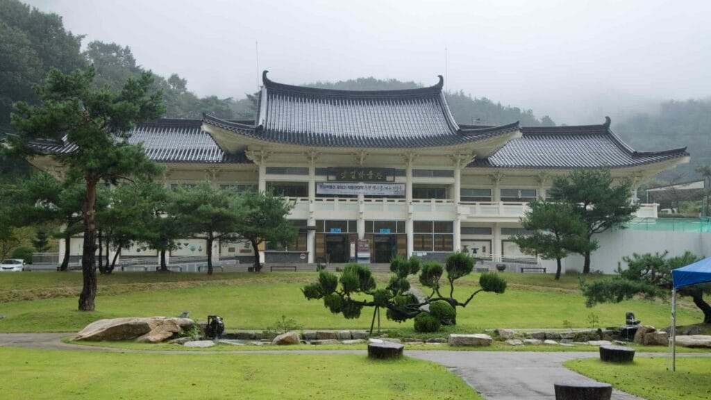 The Mungyeong Museum stands across a green lawn in light rain, its tiled roof and colonnade reflected on wet paving stones.