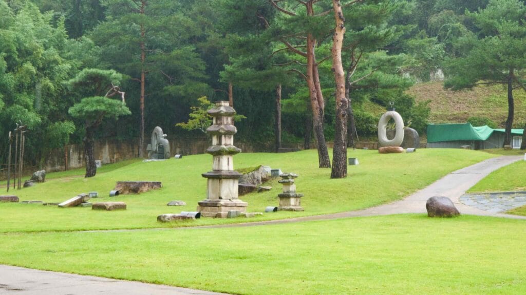 Stone pagodas and modern sculptures dot a grassy garden at the museum grounds in Mungyeong, with pine trees and walkways between them.