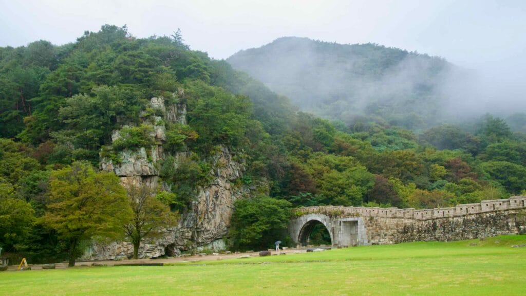 A stone arch bridge hugs the cliff inside the Saejae fortress valley.