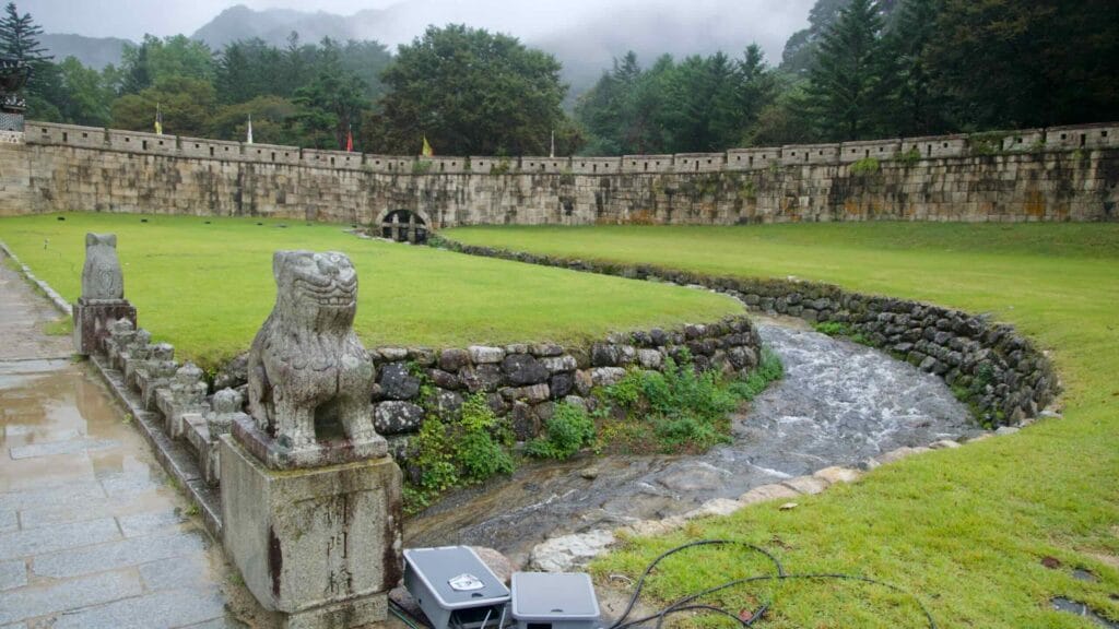 Stone guardian beasts flank a rain-wet path beside a stream and old fortress wall.