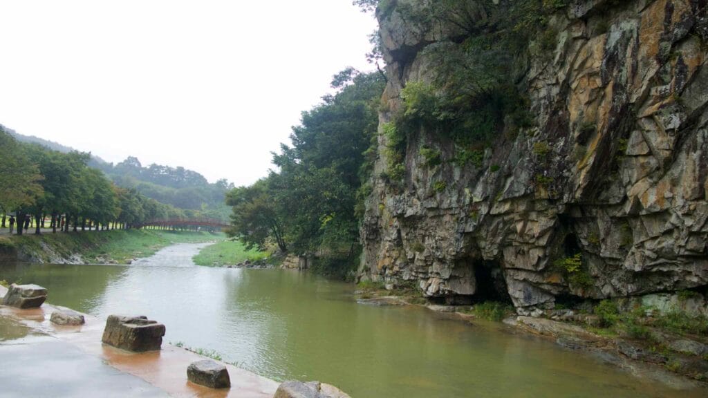 A rocky cliff and river bend frame a tree-lined promenade and footbridge in the valley.