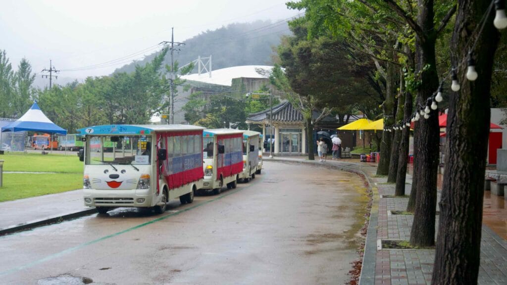 A tourist road train waits under dripping trees near the entrance on a rainy afternoon.