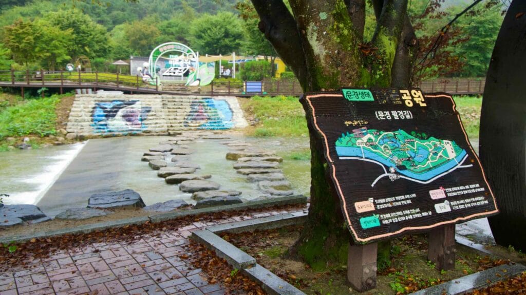 Stepping stones cross below a weir beside a carved park map sign under the trees.