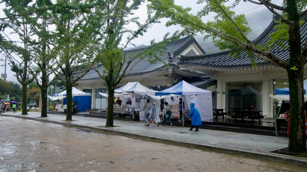 Festival stalls and visitors in rain gear line the covered sidewalk near the entrance.
