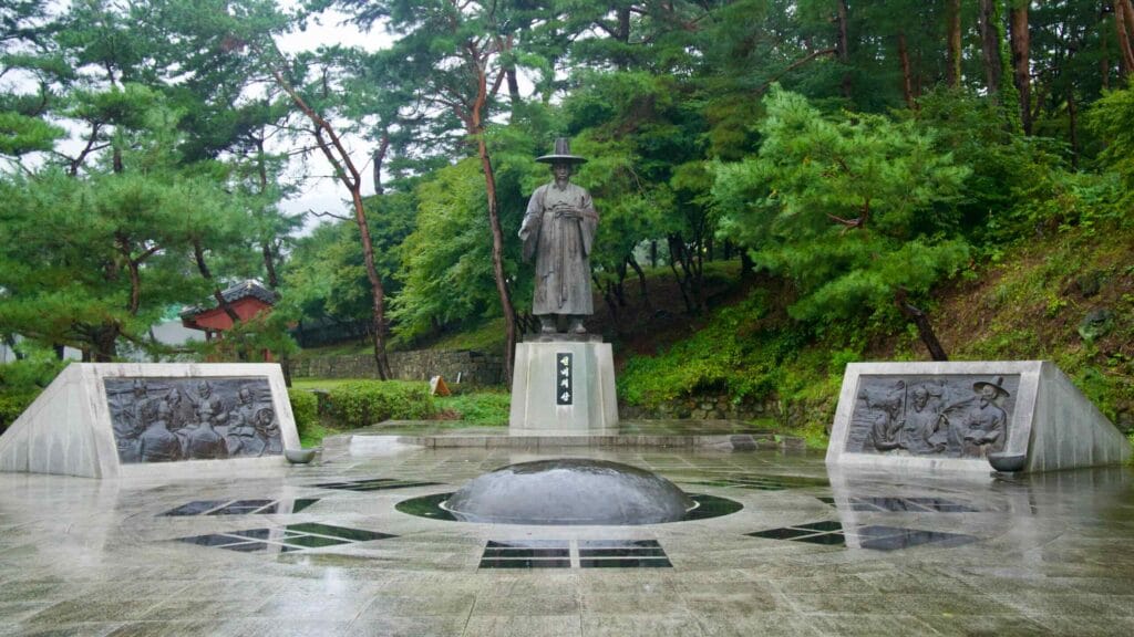 A quiet memorial square holds a statue and reliefs after rain at Mungyeong Saejae.