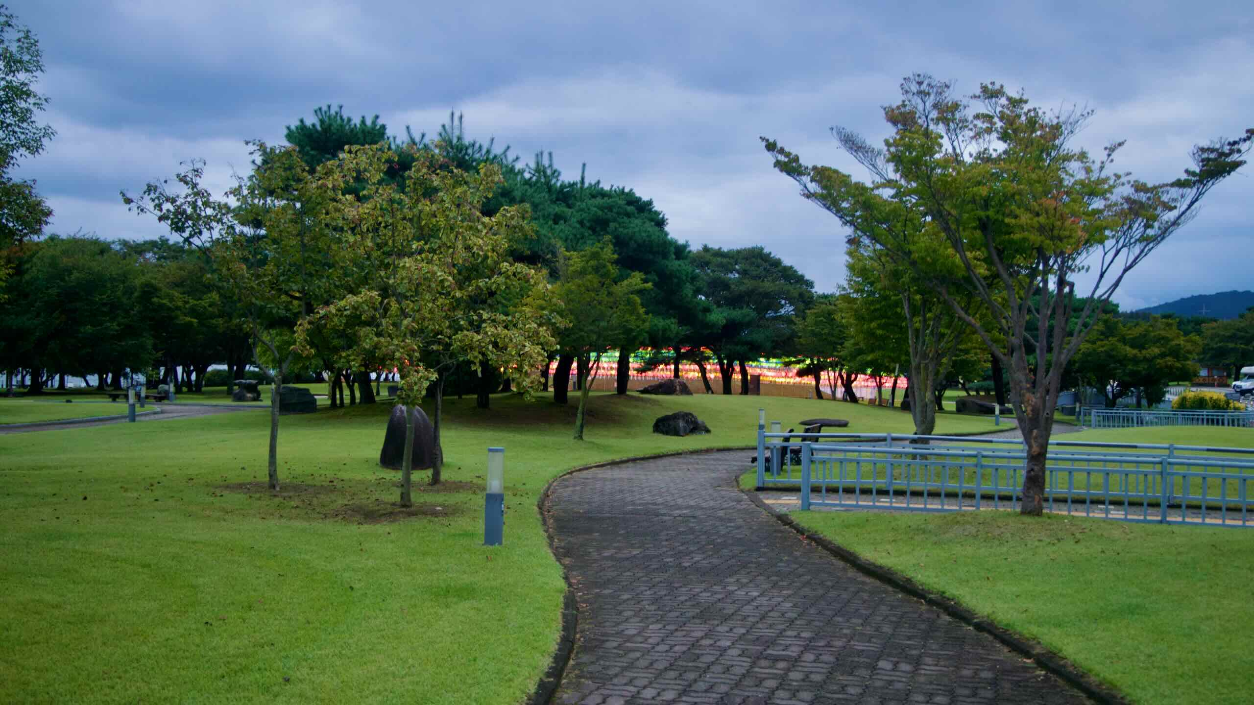 A curving brick path crosses Jungangtap Park with the Rainbow Bridge glowing in the distance.