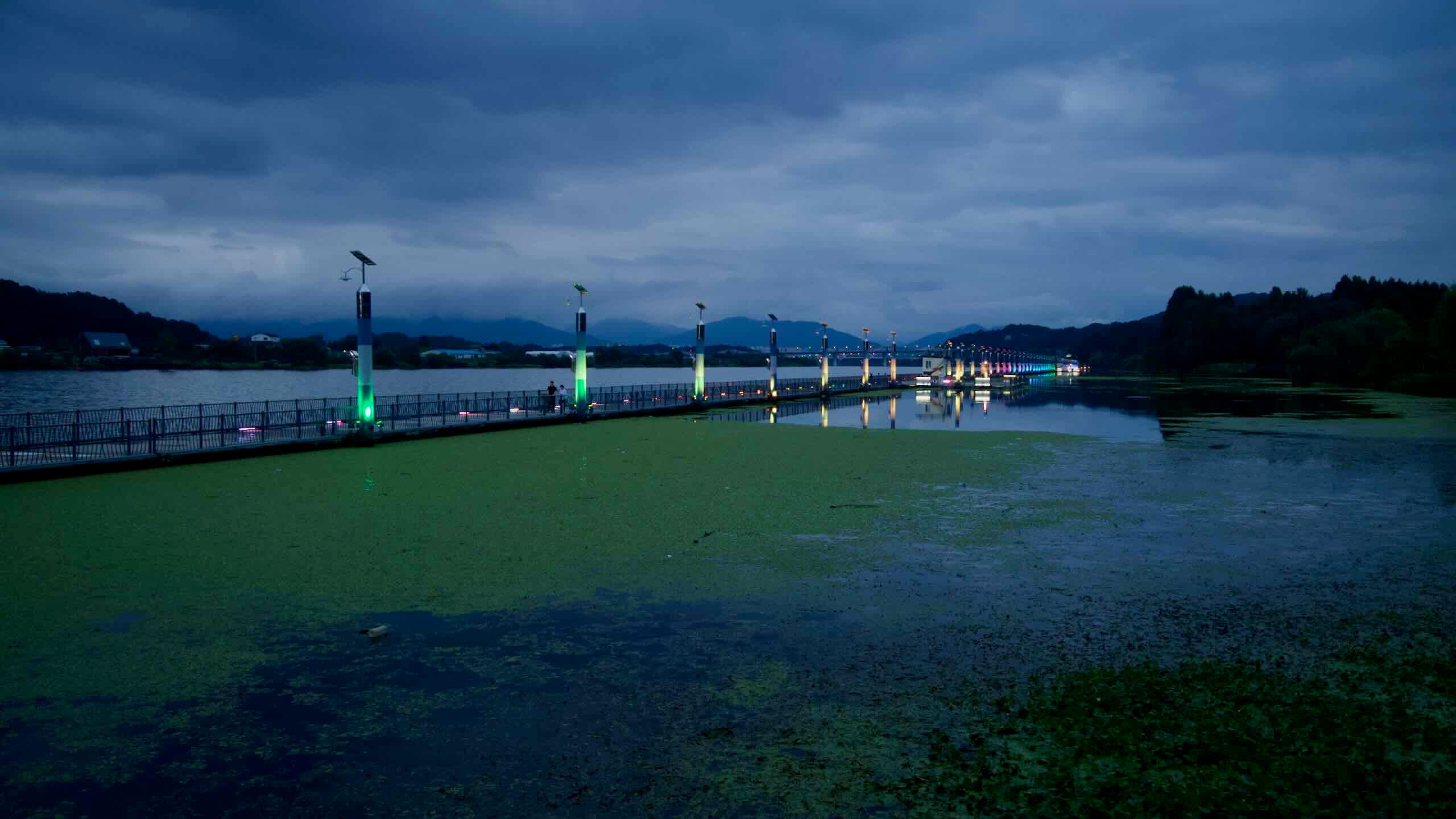 A long line of lighted pylons guides the floating pedestrian bridge across the water near Jungangtap Park.