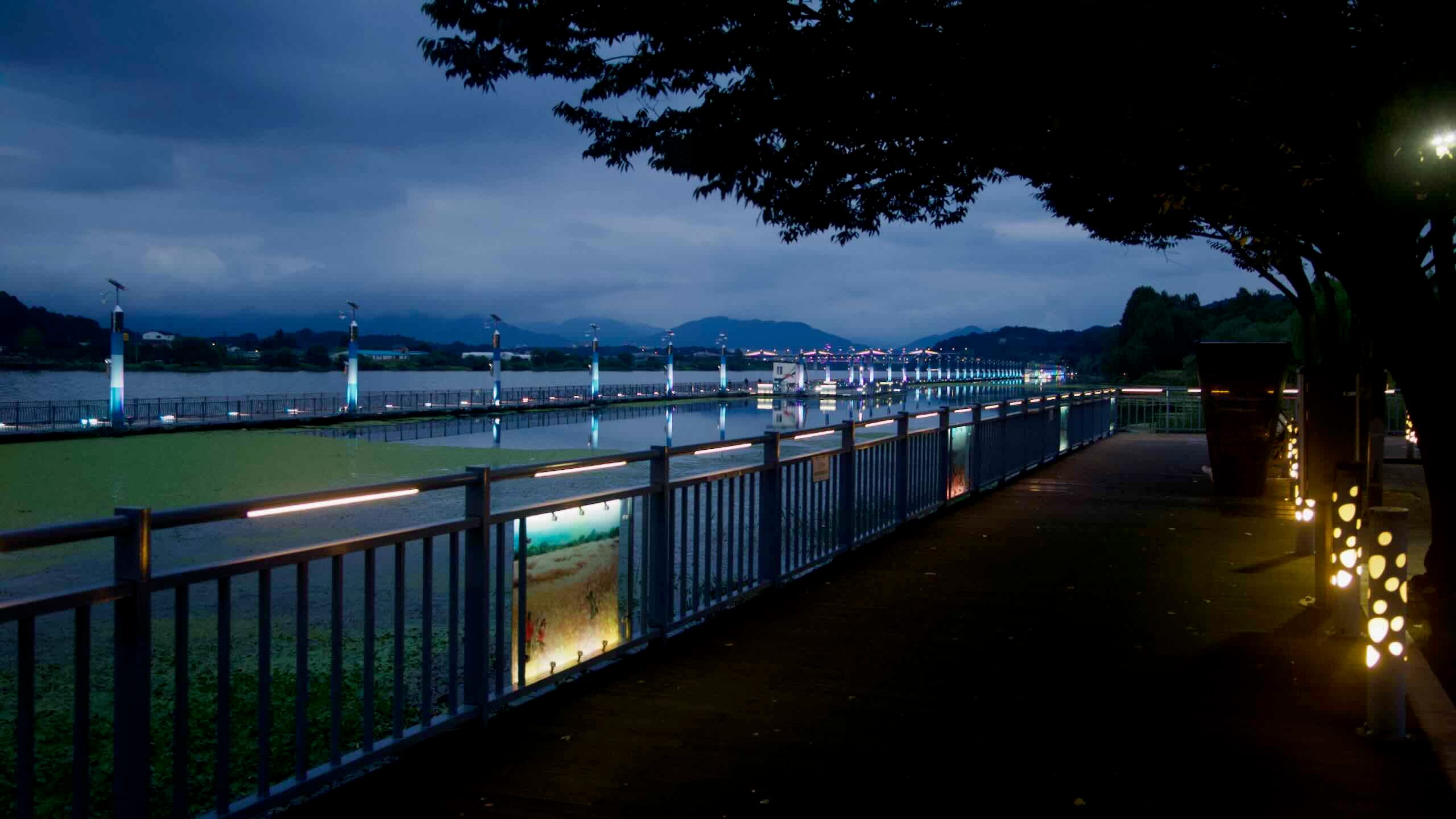 A riverside promenade with railings and lamp bollards parallels the Rainbow Bridge at Jungangtap Park.