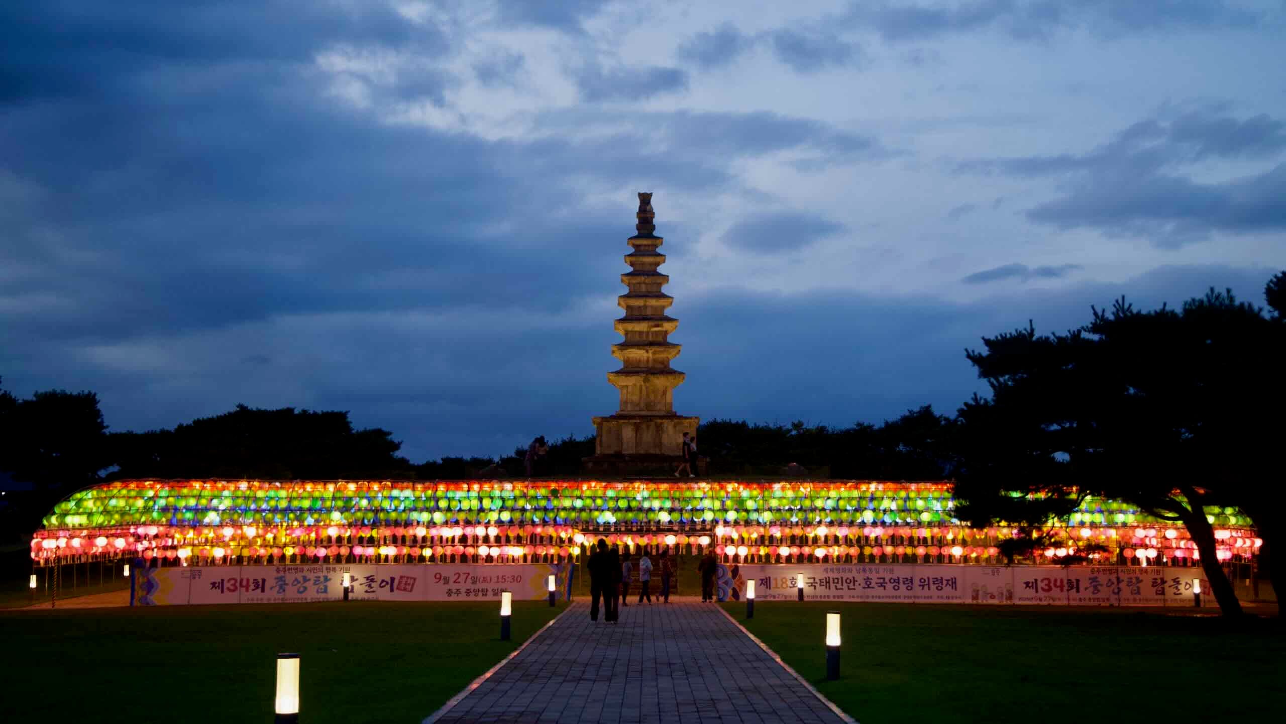 A straight brick path leads under glowing lanterns toward Chungju’s seven-story Central Tower.