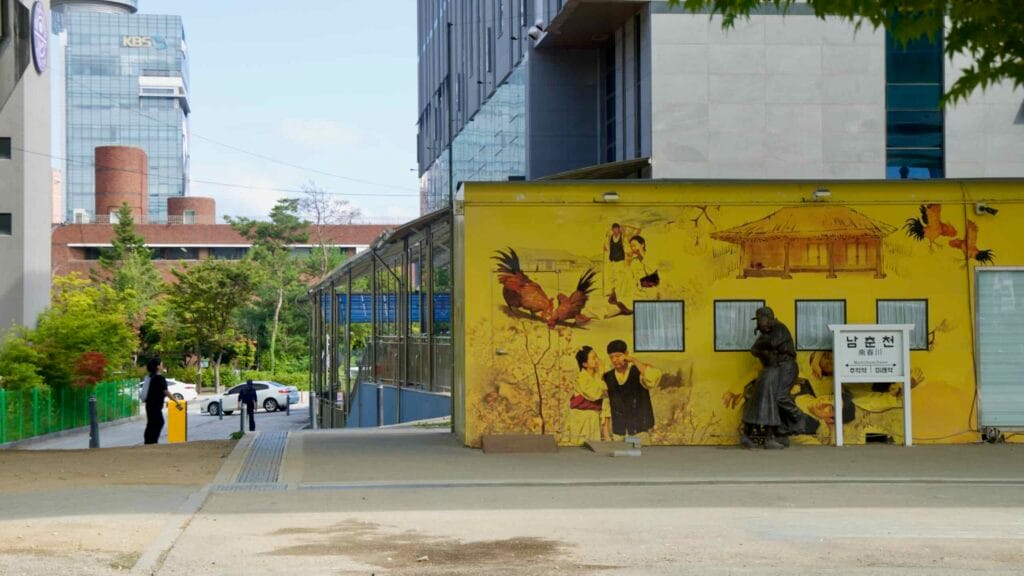 A yellow village-scene mural and street statue stand near the KBS tower in Chuncheon.