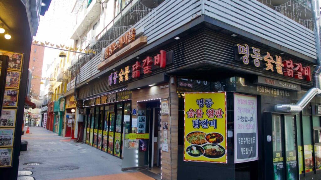 A corner dakgalbi restaurant covered in menu boards and signs stands at the junction of two narrow lanes.