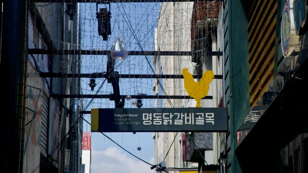 An overhead sign reading Dakgalbi Street with a yellow rooster icon hangs above the restaurant lane in Myeongdong.