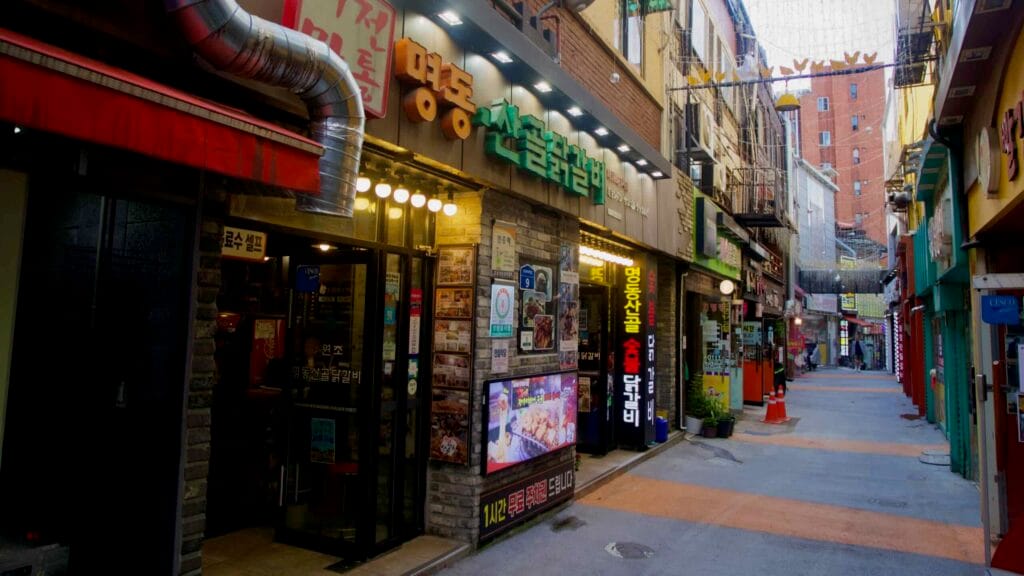 A row of dakgalbi restaurants faces a narrow pedestrian lane lit by string lights and colorful façades.